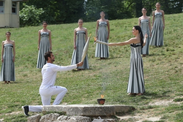 扮演最高女祭司的演員瑪麗·米娜（前右）點(diǎn)燃第一棒火炬手——希臘運(yùn)動員、東京奧運(yùn)會男子賽艇項目金牌得主斯特凡諾斯·恩圖斯科斯手中的火炬。新華社記者 趙丁喆 攝