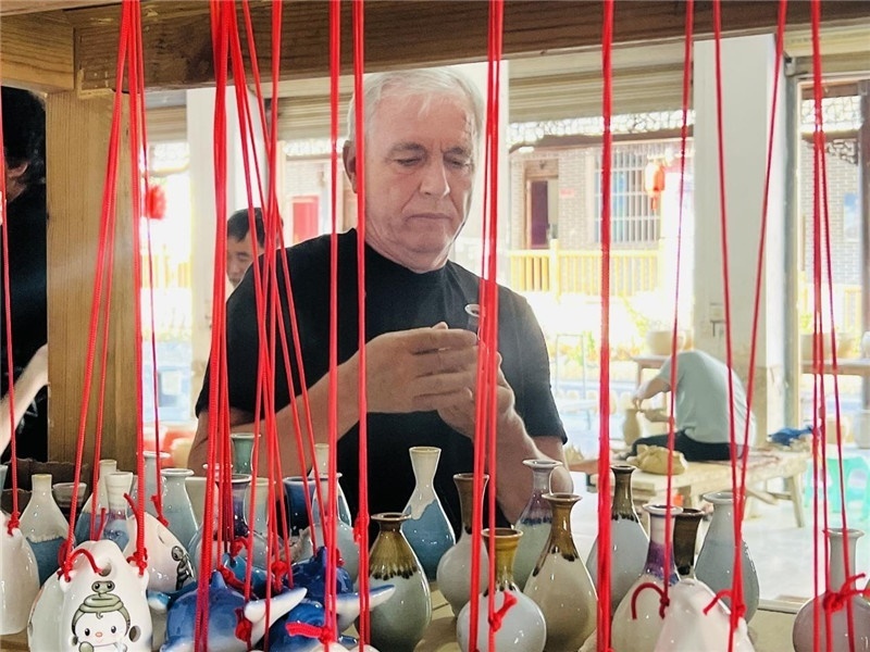 鄉(xiāng)愁如同一根根紅線，將游子與故鄉(xiāng)永遠地牽連到一起?；逄账囄幕瘎?chuàng)意一條街的母氏陶藝館內，紅線吊著陶瓶，思緒游走萬里。摩爾多瓦“信息標簽”通訊社社長亞歷山大·塔納斯端詳著館內展示的陶藝作品，并挑選出心儀的小擺件帶回摩爾多瓦，也帶走花茂村的“鄉(xiāng)愁”。（張亞欣攝）